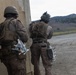 Two military personnel in full combat gear are positioned against a concrete wall, appearing to conduct a tactical exercise or training operation. Both soldiers are wearing desert camouflage uniforms, tactical vests, and helmets, and are carrying what appear to be small drone components or similar equipment attached to their gear. The setting appears to be a training range or mock urban environment with hills visible in the background.