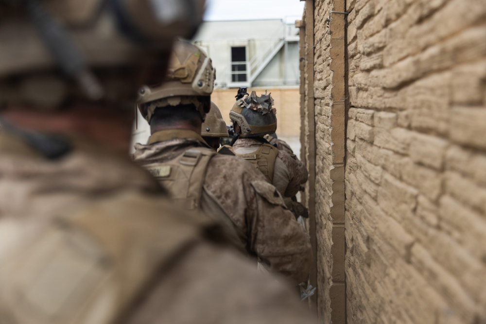 The image shows military personnel in full combat gear moving along a narrow passage between a brick wall and a building. The soldiers are wearing tan/desert camouflage uniforms and tactical helmets with mounted equipment. The perspective is taken from behind the group, suggesting a stacked formation commonly used in urban operations.