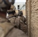 The image shows military personnel in full combat gear moving along a narrow passage between a brick wall and a building. The soldiers are wearing tan/desert camouflage uniforms and tactical helmets with mounted equipment. The perspective is taken from behind the group, suggesting a stacked formation commonly used in urban operations.