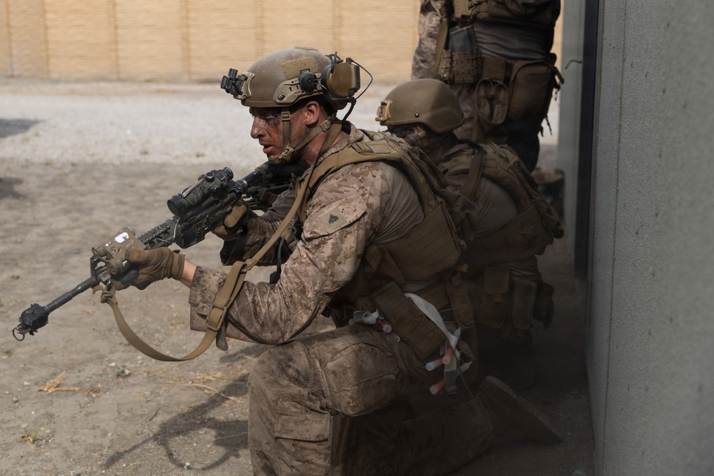 The image shows a group of military personnel in full combat gear conducting what appears to be a tactical building entry or clearing operation. The lead soldier is aiming a rifle with an optical scope while stacked against a wall near a doorway. The personnel are wearing desert camouflage uniforms consistent with U.S. Marine Corps MARPAT pattern.