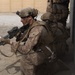 The image shows a group of military personnel in full combat gear conducting what appears to be a tactical building entry or clearing operation. The lead soldier is aiming a rifle with an optical scope while stacked against a wall near a doorway. The personnel are wearing desert camouflage uniforms consistent with U.S. Marine Corps MARPAT pattern.