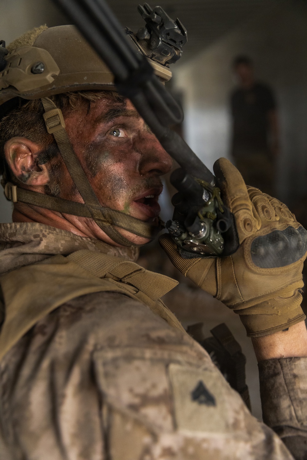 A close-up photograph of a male military personnel, likely a U.S. Marine based on visible MARPAT camouflage uniform and rank insignia, with face camouflage paint applied. He is holding a radio handset to his mouth and appears to be communicating, wearing tactical gear including a helmet with mounted accessories. His expression is intense and his face shows signs of exertion or stress.