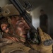 A close-up photograph of a male military personnel, likely a U.S. Marine based on visible MARPAT camouflage uniform and rank insignia, with face camouflage paint applied. He is holding a radio handset to his mouth and appears to be communicating, wearing tactical gear including a helmet with mounted accessories. His expression is intense and his face shows signs of exertion or stress.