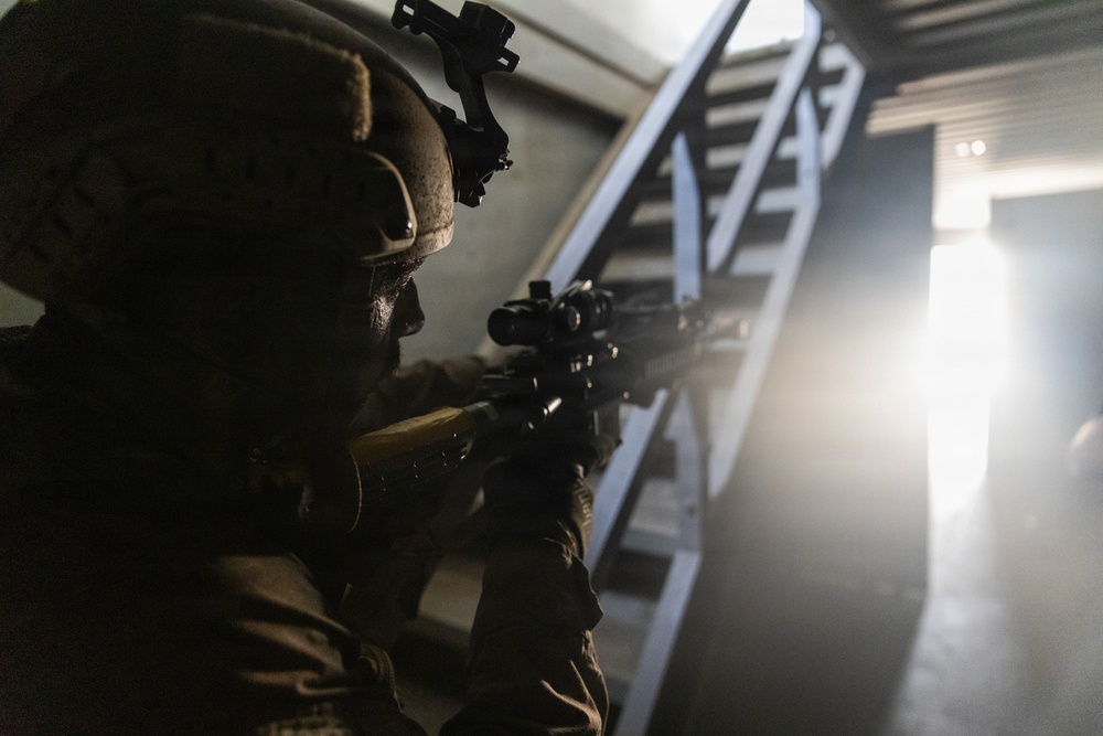 A heavily equipped soldier or tactical operator is seen in a low-light indoor environment, aiming a rifle with an optical scope toward a stairwell area. The scene is dramatically lit with a bright light source in the background, creating strong contrast and silhouetting effects. The setting appears to be a building clearance or tactical training exercise.