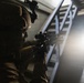 A heavily equipped soldier or tactical operator is seen in a low-light indoor environment, aiming a rifle with an optical scope toward a stairwell area. The scene is dramatically lit with a bright light source in the background, creating strong contrast and silhouetting effects. The setting appears to be a building clearance or tactical training exercise.