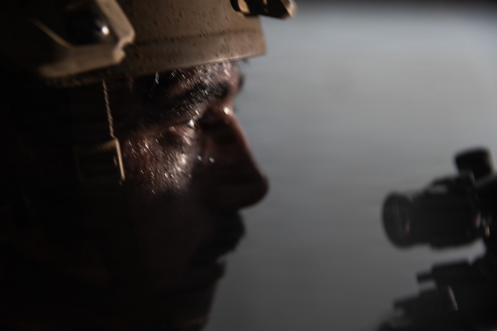 A close-up, dramatically lit photograph of a person wearing a military-style helmet, shown in profile. The image is dark and moody, with selective lighting illuminating portions of the subject's face. A weapon scope or optical device is partially visible in the lower right of the frame.