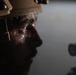 A close-up, dramatically lit photograph of a person wearing a military-style helmet, shown in profile. The image is dark and moody, with selective lighting illuminating portions of the subject's face. A weapon scope or optical device is partially visible in the lower right of the frame.