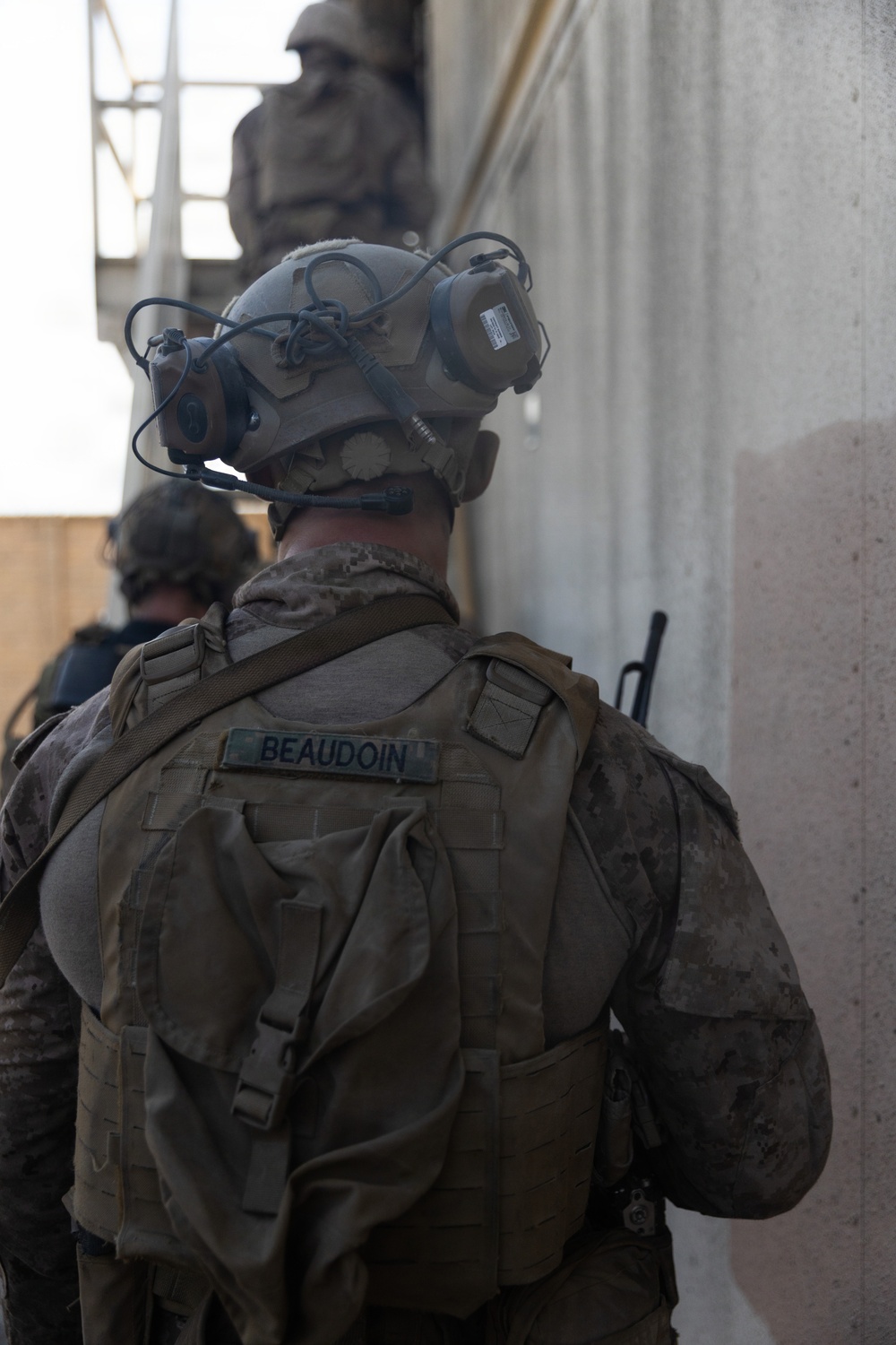 A military service member identified by the name tape 'BEAUDOIN' on their tactical vest is photographed from behind during what appears to be a training exercise or operational movement. The individual is wearing Marine Corps digital camouflage (MARPAT), a combat helmet with communications equipment, and a tactical plate carrier. Additional personnel are visible in the background near a concrete structure.