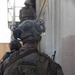 A military service member identified by the name tape 'BEAUDOIN' on their tactical vest is photographed from behind during what appears to be a training exercise or operational movement. The individual is wearing Marine Corps digital camouflage (MARPAT), a combat helmet with communications equipment, and a tactical plate carrier. Additional personnel are visible in the background near a concrete structure.
