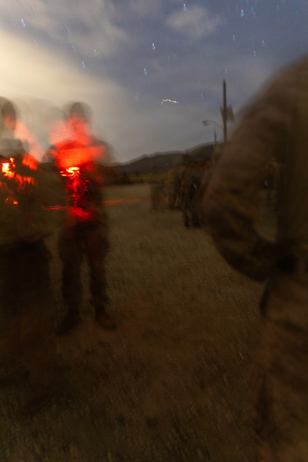 A heavily motion-blurred photograph taken at dusk or dawn showing multiple figures in what appears to be military or tactical gear on a dirt road or path. A prominent red light source (possibly a flare, signal light, or red-filtered flashlight) illuminates figures on the left side of the frame. The image exhibits significant camera motion blur, suggesting a long exposure or movement during capture.