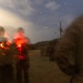 A heavily motion-blurred photograph taken at dusk or dawn showing multiple figures in what appears to be military or tactical gear on a dirt road or path. A prominent red light source (possibly a flare, signal light, or red-filtered flashlight) illuminates figures on the left side of the frame. The image exhibits significant camera motion blur, suggesting a long exposure or movement during capture.