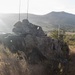 A military armored vehicle is positioned on a hillside in a semi-arid landscape, partially concealed with camouflage netting and natural vegetation. The vehicle appears to be in a tactical hide or observation position, with at least one soldier visible nearby. The scene suggests a military training exercise or field operation in a rugged, mountainous terrain.
