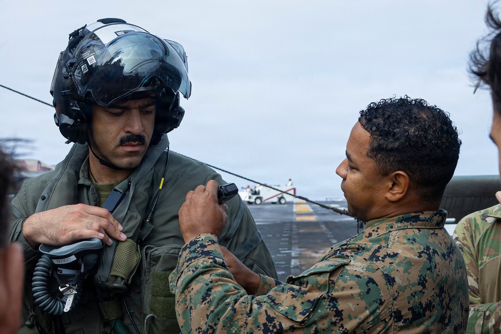 Two military personnel are on what appears to be a naval vessel flight deck. A pilot wearing a flight helmet with a dark visor and olive flight suit is being assisted or inspected by a service member in MARPAT digital camouflage uniform. The background shows flight deck markings and what appears to be ground support equipment.