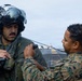 Two military personnel are on what appears to be a naval vessel flight deck. A pilot wearing a flight helmet with a dark visor and olive flight suit is being assisted or inspected by a service member in MARPAT digital camouflage uniform. The background shows flight deck markings and what appears to be ground support equipment.
