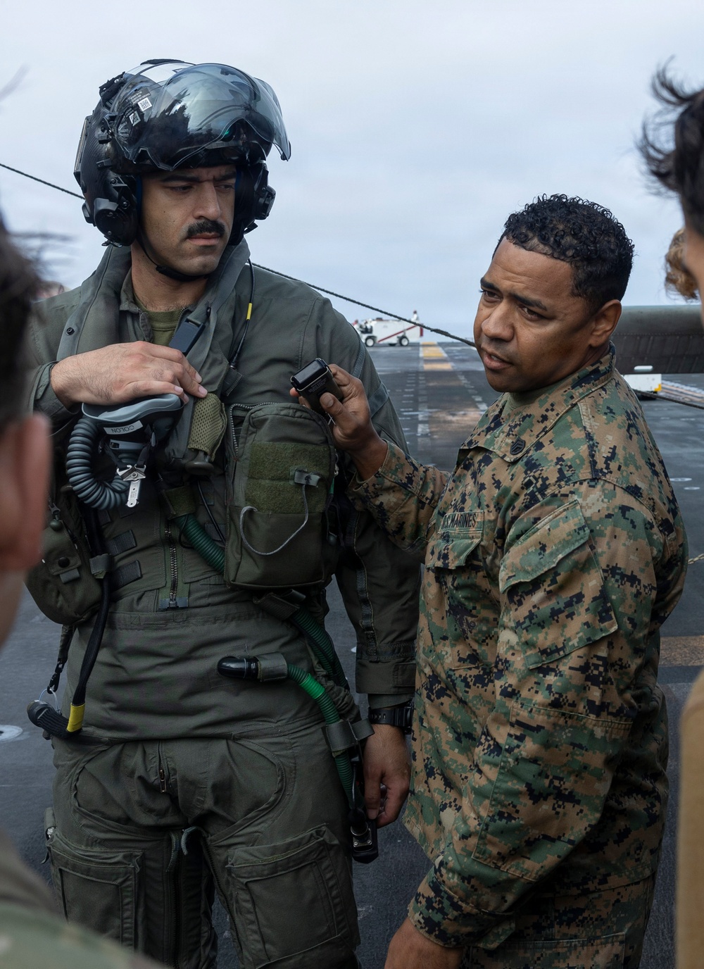 Two military personnel are on what appears to be a ship's flight deck. A pilot in full flight gear including a helmet with visor is being assisted or inspected by a U.S. Marines service member in MARPAT camouflage uniform. The Marine appears to be checking or adjusting equipment on the pilot's flight suit.