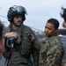 Two military personnel are on what appears to be a ship's flight deck. A pilot in full flight gear including a helmet with visor is being assisted or inspected by a U.S. Marines service member in MARPAT camouflage uniform. The Marine appears to be checking or adjusting equipment on the pilot's flight suit.