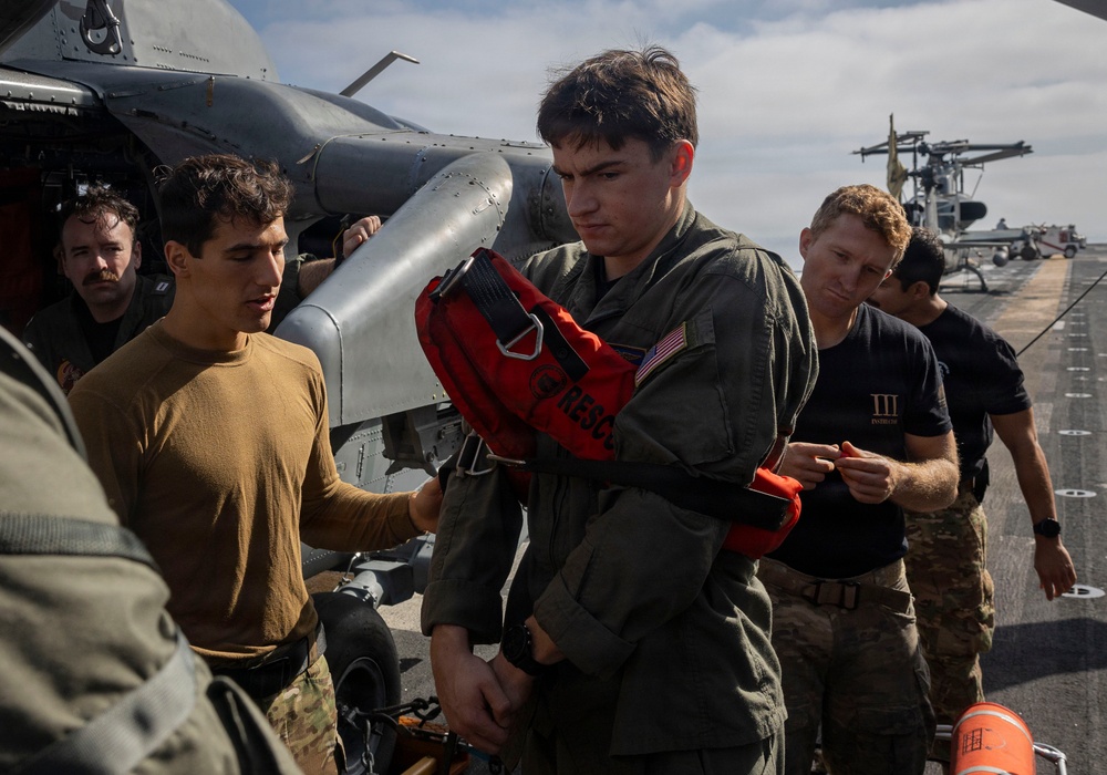 Military personnel are gathered on what appears to be a naval vessel's flight deck near a helicopter. A central figure in an olive green flight suit is holding or donning a red rescue/survival vest marked 'RESCUE' with a US flag patch visible on the sleeve. Several other personnel in various military uniforms are present around him, with additional aircraft visible in the background.