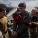 Military personnel are gathered on what appears to be a naval vessel's flight deck near a helicopter. A central figure in an olive green flight suit is holding or donning a red rescue/survival vest marked 'RESCUE' with a US flag patch visible on the sleeve. Several other personnel in various military uniforms are present around him, with additional aircraft visible in the background.