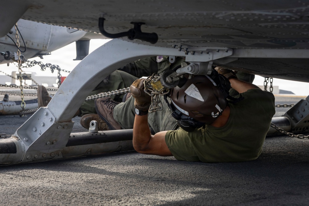 A military aviation technician or deck crew member is lying on their back beneath an aircraft, working on securing or inspecting a tie-down chain connection point on the underside of the fuselage. The setting appears to be a naval vessel flight deck or military airfield, indicated by the non-skid deck surface and multiple tie-down chains visible in the background. The worker is wearing standard flight deck personal protective equipment.