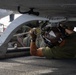 A military aviation technician or deck crew member is lying on their back beneath an aircraft, working on securing or inspecting a tie-down chain connection point on the underside of the fuselage. The setting appears to be a naval vessel flight deck or military airfield, indicated by the non-skid deck surface and multiple tie-down chains visible in the background. The worker is wearing standard flight deck personal protective equipment.