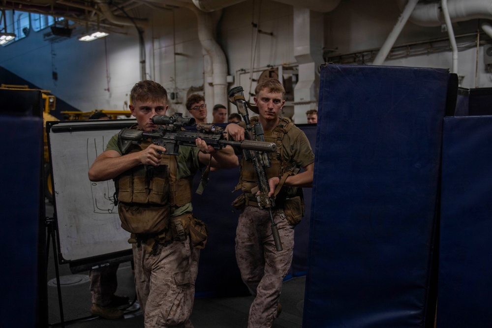 Two military personnel in combat gear and camouflage uniforms are conducting a close-quarters battle (CQB) training exercise in what appears to be the interior of a naval vessel. They are moving in a tactical formation around a blue padded barrier while holding assault rifles with optics. Additional personnel are visible in the background observing the drill.