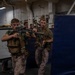 Two military personnel in combat gear and camouflage uniforms are conducting a close-quarters battle (CQB) training exercise in what appears to be the interior of a naval vessel. They are moving in a tactical formation around a blue padded barrier while holding assault rifles with optics. Additional personnel are visible in the background observing the drill.