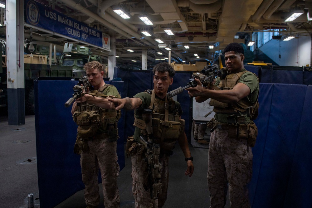 Three U.S. Marines in full combat gear are conducting a training exercise aboard the USS Makin Island (LHD-8), a U.S. Navy amphibious assault ship. They are in the ship's well deck or vehicle bay, aiming rifles in a tactical formation. The ship's name and hull designation are clearly visible on a sign in the background.