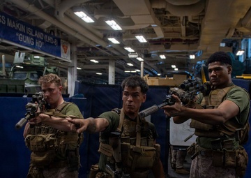 Three U.S. Marines in full combat gear are conducting a training exercise aboard the USS Makin Island (LHD-8), a U.S. Navy amphibious assault ship. They are in the ship's well deck or vehicle bay, aiming rifles in a tactical formation. The ship's name and hull designation are clearly visible on a sign in the background.