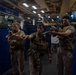 Three U.S. Marines in full combat gear are conducting a training exercise aboard the USS Makin Island (LHD-8), a U.S. Navy amphibious assault ship. They are in the ship's well deck or vehicle bay, aiming rifles in a tactical formation. The ship's name and hull designation are clearly visible on a sign in the background.