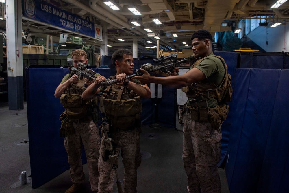 Three U.S. Marines in combat gear are conducting weapons training or instruction aboard the USS Makin Island (LHD-8), a Wasp-class amphibious assault ship. The Marines are wearing MARPAT camouflage uniforms and tactical vests while handling rifles, with one Marine appearing to instruct the others on weapon handling techniques. The setting is the ship's well deck or vehicle storage bay, identifiable by the ship's name banner and military vehicles visible in the background.