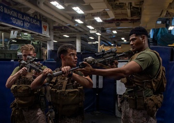 Three U.S. Marines in combat gear are conducting weapons training or instruction aboard the USS Makin Island (LHD-8), a Wasp-class amphibious assault ship. The Marines are wearing MARPAT camouflage uniforms and tactical vests while handling rifles, with one Marine appearing to instruct the others on weapon handling techniques. The setting is the ship's well deck or vehicle storage bay, identifiable by the ship's name banner and military vehicles visible in the background.
