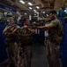 Three U.S. Marines in combat gear are conducting weapons training or instruction aboard the USS Makin Island (LHD-8), a Wasp-class amphibious assault ship. The Marines are wearing MARPAT camouflage uniforms and tactical vests while handling rifles, with one Marine appearing to instruct the others on weapon handling techniques. The setting is the ship's well deck or vehicle storage bay, identifiable by the ship's name banner and military vehicles visible in the background.