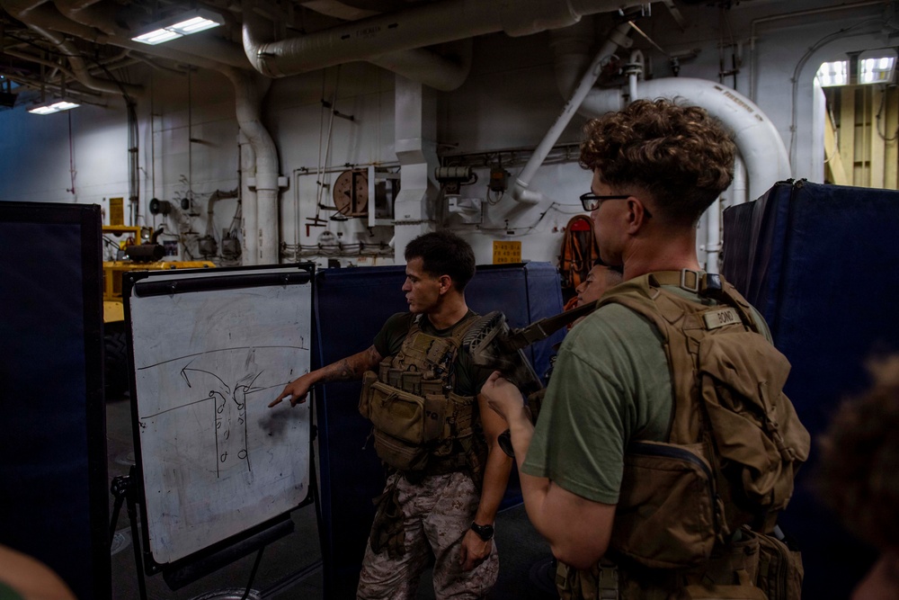 Two military personnel in tactical gear are engaged in a briefing session aboard what appears to be a naval vessel. One individual in camouflage uniform is pointing at a whiteboard displaying a tactical diagram with arrows and lines. The setting is an interior compartment of a ship, identifiable by exposed pipes, industrial fittings, and overhead lighting.