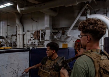 Two military personnel in tactical gear are engaged in a briefing session aboard what appears to be a naval vessel. One individual in camouflage uniform is pointing at a whiteboard displaying a tactical diagram with arrows and lines. The setting is an interior compartment of a ship, identifiable by exposed pipes, industrial fittings, and overhead lighting.
