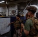 Two military personnel in tactical gear are engaged in a briefing session aboard what appears to be a naval vessel. One individual in camouflage uniform is pointing at a whiteboard displaying a tactical diagram with arrows and lines. The setting is an interior compartment of a ship, identifiable by exposed pipes, industrial fittings, and overhead lighting.