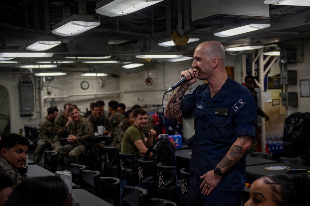 A U.S. Navy sailor in dark blue uniform is speaking into a microphone aboard what appears to be a naval vessel, addressing a seated audience of military personnel. The speaker is bald with visible tattoos on both arms and is wearing a name tag. The audience, consisting of sailors and Marines in various uniforms, appears engaged and amused.