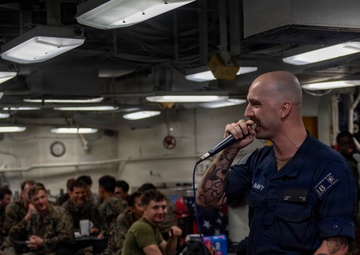 A U.S. Navy sailor in dark blue uniform is speaking into a microphone aboard what appears to be a naval vessel, addressing a seated audience of military personnel. The speaker is bald with visible tattoos on both arms and is wearing a name tag. The audience, consisting of sailors and Marines in various uniforms, appears engaged and amused.