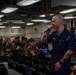 A U.S. Navy sailor in dark blue uniform is speaking into a microphone aboard what appears to be a naval vessel, addressing a seated audience of military personnel. The speaker is bald with visible tattoos on both arms and is wearing a name tag. The audience, consisting of sailors and Marines in various uniforms, appears engaged and amused.