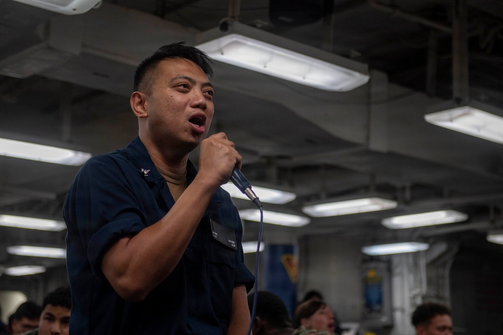 A person wearing a dark navy blue military uniform is speaking or singing into a handheld microphone in what appears to be the interior of a naval vessel. The individual has a name tag on their uniform and appears to be addressing or performing for a group of people visible in the background. The setting is an enclosed, industrial-style space with fluorescent lighting mounted on a low ceiling.