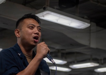 A person wearing a dark navy blue military uniform is speaking or singing into a handheld microphone in what appears to be the interior of a naval vessel. The individual has a name tag on their uniform and appears to be addressing or performing for a group of people visible in the background. The setting is an enclosed, industrial-style space with fluorescent lighting mounted on a low ceiling.