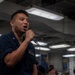 A person wearing a dark navy blue military uniform is speaking or singing into a handheld microphone in what appears to be the interior of a naval vessel. The individual has a name tag on their uniform and appears to be addressing or performing for a group of people visible in the background. The setting is an enclosed, industrial-style space with fluorescent lighting mounted on a low ceiling.