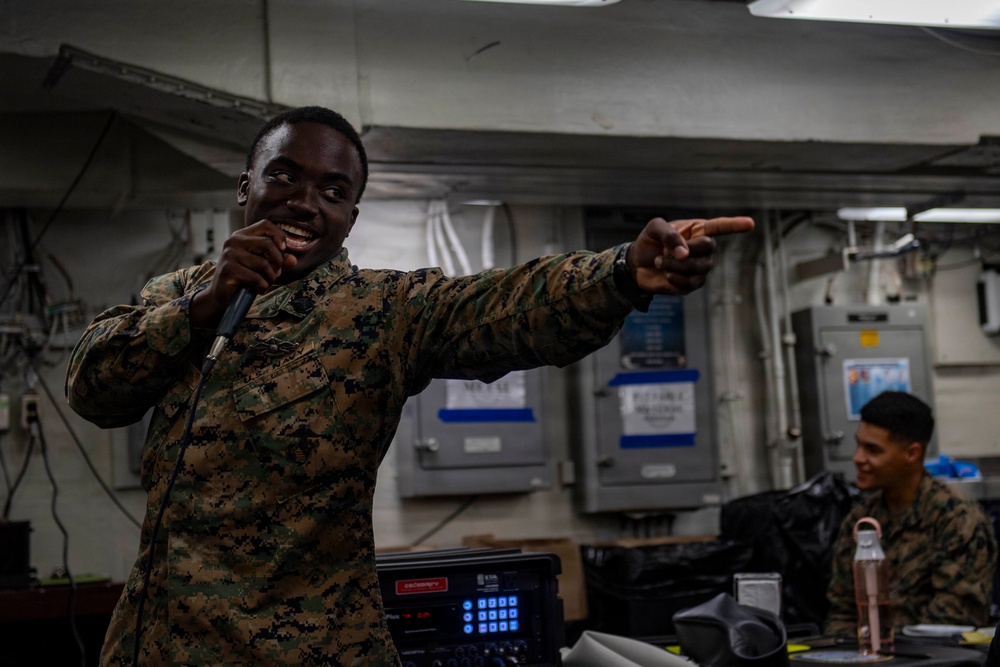 A U.S. Marine in digital camouflage (MARPAT) uniform is holding a microphone and pointing outward, appearing to be performing or emceeing an event aboard what appears to be a naval vessel. A second Marine is visible in the background, smiling. The setting appears to be an interior compartment of a ship with visible electrical panels, cables, and industrial infrastructure.