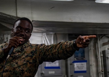 A U.S. Marine in digital camouflage (MARPAT) uniform is holding a microphone and pointing outward, appearing to be performing or emceeing an event aboard what appears to be a naval vessel. A second Marine is visible in the background, smiling. The setting appears to be an interior compartment of a ship with visible electrical panels, cables, and industrial infrastructure.