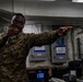 A U.S. Marine in digital camouflage (MARPAT) uniform is holding a microphone and pointing outward, appearing to be performing or emceeing an event aboard what appears to be a naval vessel. A second Marine is visible in the background, smiling. The setting appears to be an interior compartment of a ship with visible electrical panels, cables, and industrial infrastructure.