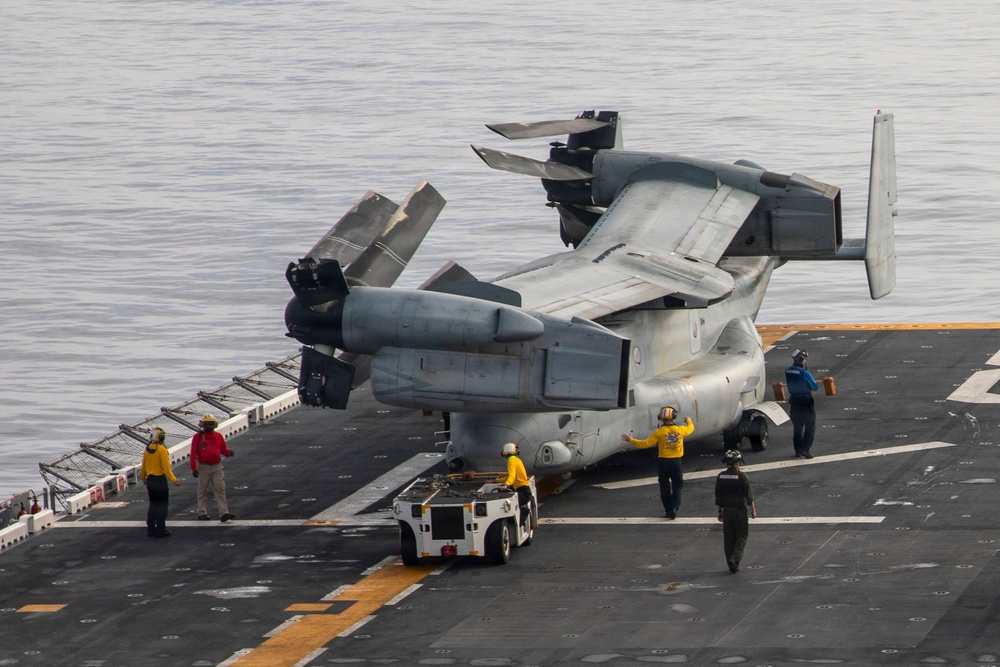 A tiltrotor aircraft, identifiable as a Bell Boeing V-22 Osprey, is being maneuvered on the flight deck of a naval vessel with its rotors folded for storage. Ground crew members in colored jerseys are guiding the aircraft as it is towed by a yellow aircraft tug. The scene takes place over open water, visible in the background beyond the ship's deck edge.