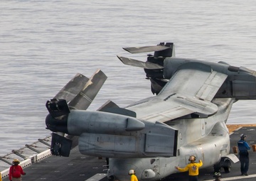 A tiltrotor aircraft, identifiable as a Bell Boeing V-22 Osprey, is being maneuvered on the flight deck of a naval vessel with its rotors folded for storage. Ground crew members in colored jerseys are guiding the aircraft as it is towed by a yellow aircraft tug. The scene takes place over open water, visible in the background beyond the ship's deck edge.