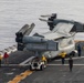 A tiltrotor aircraft, identifiable as a Bell Boeing V-22 Osprey, is being maneuvered on the flight deck of a naval vessel with its rotors folded for storage. Ground crew members in colored jerseys are guiding the aircraft as it is towed by a yellow aircraft tug. The scene takes place over open water, visible in the background beyond the ship's deck edge.