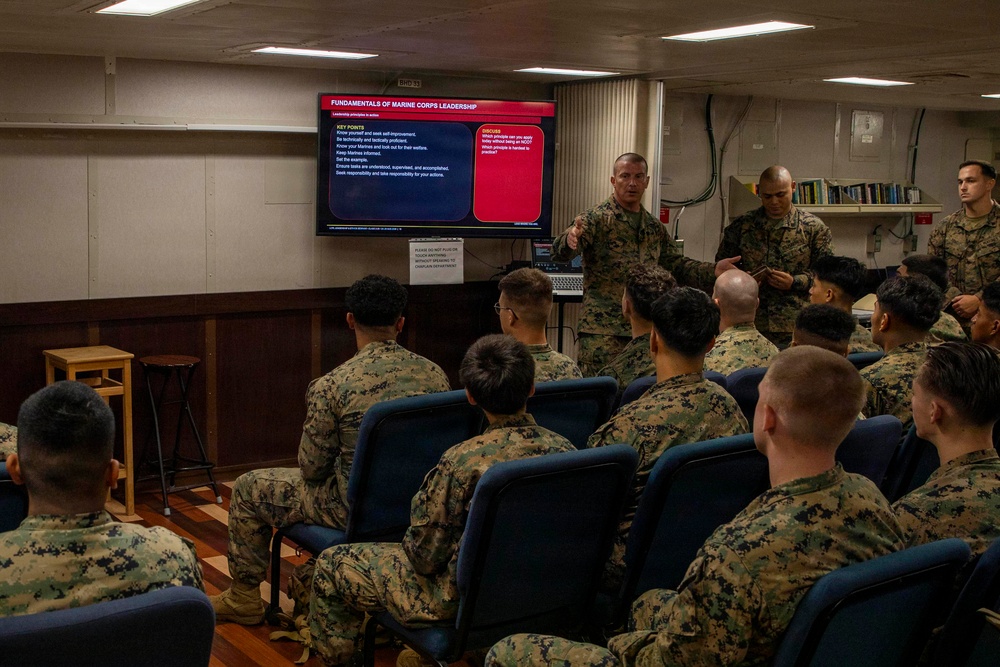 A Marine Corps instructor is conducting a leadership training class for a group of Marines seated in blue chairs. The presentation displayed on a large screen is titled 'Fundamentals of Marine Corps Leadership,' covering leadership principles in action. The setting appears to be a briefing room or classroom aboard a ship or military facility.
