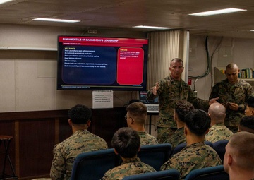 A Marine Corps instructor is conducting a leadership training class for a group of Marines seated in blue chairs. The presentation displayed on a large screen is titled 'Fundamentals of Marine Corps Leadership,' covering leadership principles in action. The setting appears to be a briefing room or classroom aboard a ship or military facility.