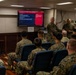 A Marine Corps instructor is conducting a leadership training class for a group of Marines seated in blue chairs. The presentation displayed on a large screen is titled 'Fundamentals of Marine Corps Leadership,' covering leadership principles in action. The setting appears to be a briefing room or classroom aboard a ship or military facility.